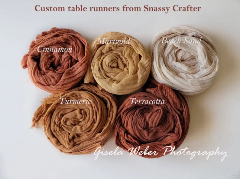 May include: Six rolled fabric table runners in various shades of brown, including cinnamon, marigold, beach sand, turmeric, terracotta, and a lighter brown. The fabric is a soft, gauzy material. The text "Custom table runners from Snassy Crafter" and "Gisela Weber Photography" is visible in the image.
