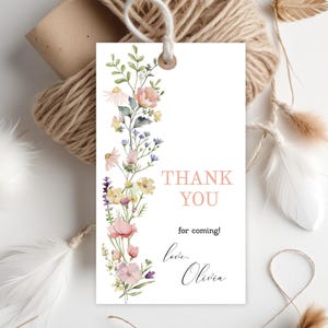 May include: A white gift tag with a floral watercolor design and the text "THANK YOU for coming! love, Olivia." The tag is decorated with pink, yellow, and purple flowers and green leaves. A spool of twine and feathers are in the background.