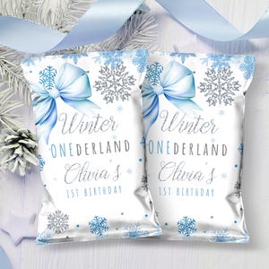 May include: Two white treat bags with a winter theme. Each bag features a light blue bow, snowflakes, and the text "Winter Onederland Olivia's 1st Birthday". Decorative elements include light blue ribbon and pine branches.