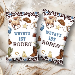 May include: Two white party favor bags with a cowboy theme. Each bag features a running horse, cowboy hat, and the text "WAYAT'S 1ST RODEO" in blue. The bags have a brown and blue cowhide pattern.