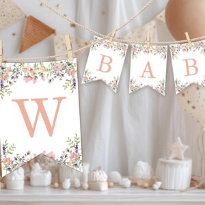 May include: A decorative banner featuring individual white pennants hanging from a rope with wooden clips. Each pennant displays a large, light orange letter and is decorated with a watercolor floral border of pink, purple, and green foliage.