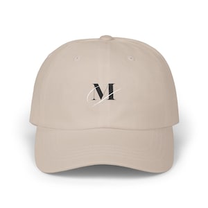 Könnte beinhalten: Beige Baseballkappe mit gebogenem Schirm. Die Kappe zeigt den schwarzen Buchstaben "M" mit einem zarten weißen Schreibschrift-Akzent. Der Hut ist aus weichem Stoff und für den lässigen Gebrauch geeignet.