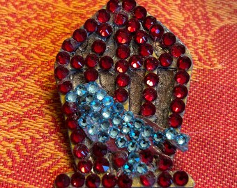 May include: A decorative birdcage pin, adorned with red and blue rhinestones. The cage is outlined in silver, with a blue bird inside. The pin is set against a red and orange patterned background.