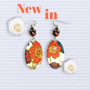 Pode incluir: Brincos ovais com um desenho floral em laranja, vermelho e amarelo. Os brincos têm uma conta e um gancho de cor bronze. As palavras "New in" estão em laranja no topo. Rosas brancas em ambos os lados.