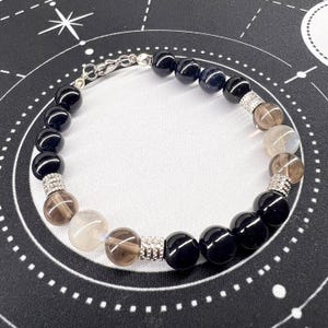 Peut inclure: Un bracelet de perles composé de perles noires, de quartz fumé et de pierres de lune irisées. Le bracelet est orné d'entretoises argentées et d'un fermoir. Les perles sont rondes et polies, créant un contraste de couleurs et de textures.