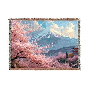 Könnte beinhalten: Gewebte Decke mit einer japanischen Landschaft, die einen schneebedeckten Berg, Kirschblütenbäume und eine Pagode zeigt. Das Design verwendet Rosa-, Blau- und Grüntöne. Die Decke hat einen Fransenrand.