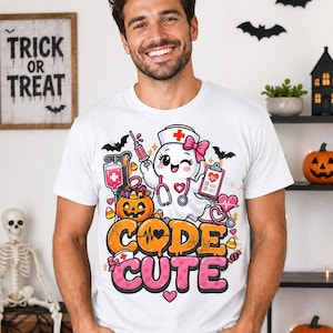 May include: A white t-shirt featuring a graphic of a cartoon ghost dressed as a nurse, holding a syringe and clipboard. The design includes a jack-o'-lantern, bats, and the text "CODE CUTE" in orange and pink bubble letters with heart accents.