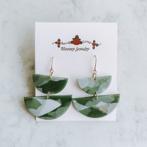 Può includere: Un paio di orecchini a forma di semicerchio sovrapposti, di colore verde e bianco con glitter dorati. Gli orecchini sono su una carta bianca con il testo "Bloomy Jewelry".