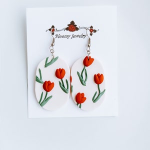 May include: A pair of white oval-shaped earrings with a red tulip floral pattern and silver hooks. The earrings are displayed on a white card with the text "Bloomy Jewelry".