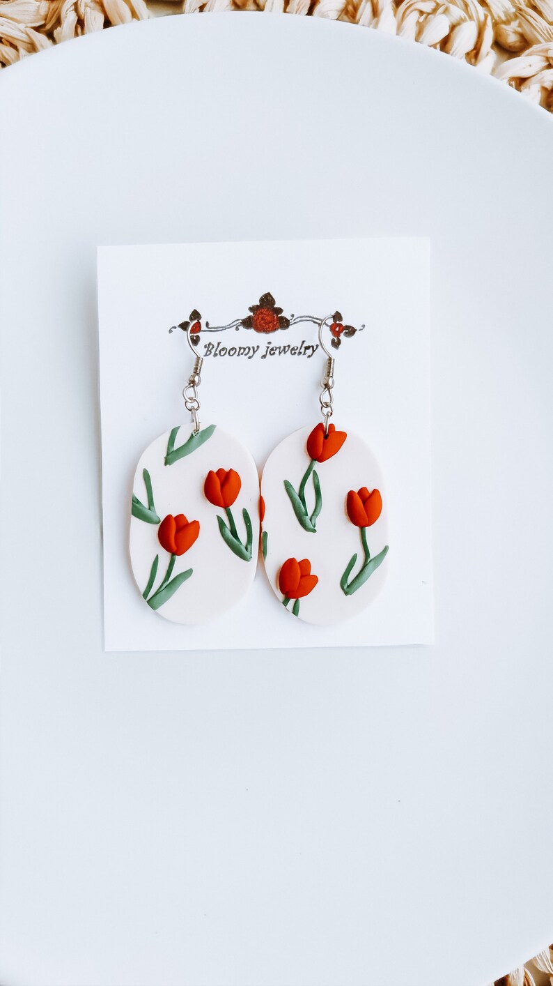 May include: A pair of white oval-shaped earrings with a red tulip floral pattern and silver hooks. The earrings are displayed on a white card with the text "Bloomy Jewelry".