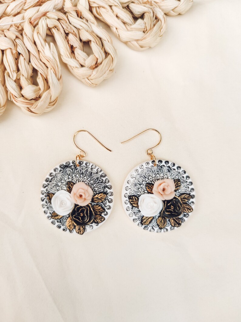K&ouml;nnte beinhalten: Ein Paar goldene Ohrringe mit wei&szlig;en und rosafarbenen Blumendesigns auf wei&szlig;em Hintergrund. Die Ohrringe sind rund und haben ein schwarzes und goldenes Mandala-Muster.