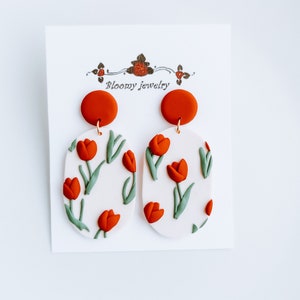 May include: A pair of white oval-shaped earrings with a red tulip pattern and a red clay top. The earrings are on a white card with the text "Bloomy Jewelry".