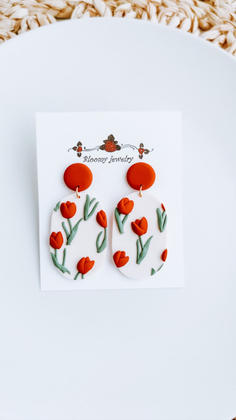 May include: A pair of white oval-shaped earrings with a red tulip pattern and a red clay top. The earrings are on a white card with the text "Bloomy Jewelry".