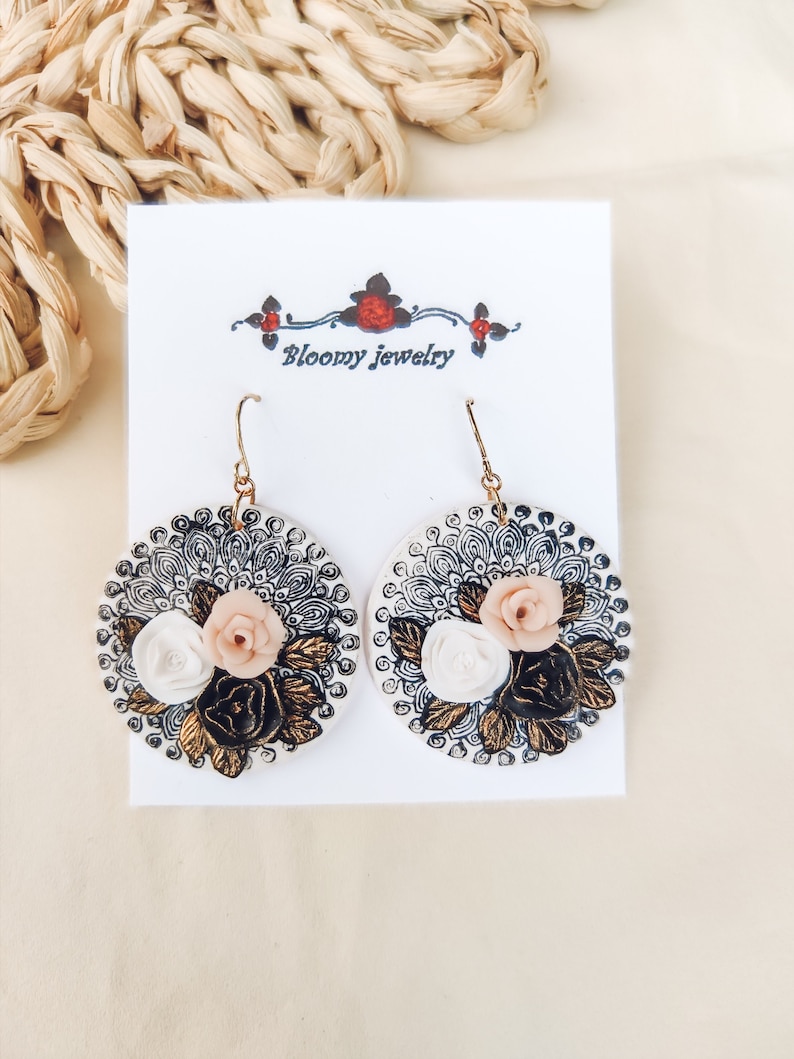 K&ouml;nnte beinhalten: Ein Paar wei&szlig;e und schwarze Blumenohrringe mit goldenen Akzenten. Die Ohrringe haben ein rundes Design mit einem schwarzen und wei&szlig;en Mandala-Muster und dreidimensionalen Blumen in Wei&szlig;, Rosa und Gold.