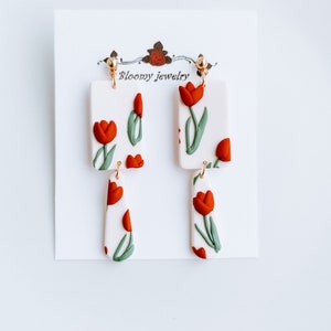 May include: A pair of dangle earrings with a white background and red and green tulip designs. The earrings are made of polymer clay and have gold-toned hooks. The earrings are labeled "Bloomy Jewelry".