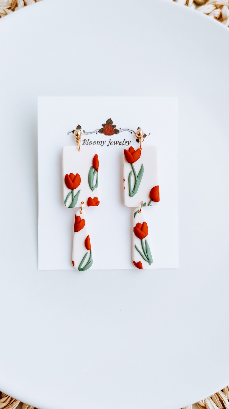 May include: A pair of dangle earrings with a white background and red and green tulip designs. The earrings are made of polymer clay and have gold-toned hooks. The earrings are labeled "Bloomy Jewelry".