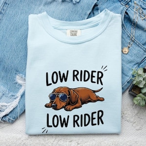 May include: A light blue t-shirt featuring a graphic of a brown dachshund wearing sunglasses. The text "LOW RIDER" is printed in black above and below the dog illustration. The shirt has a Comfort Colors brand label on the inside of the neckline.
