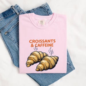 Può includere: Una t-shirt rosa chiaro con il testo arancione "CROISSANTS & CAFFEINE" sopra un'illustrazione grafica di due croissant dorati con vapore che sale, abbinata a un paio di jeans in denim blu.