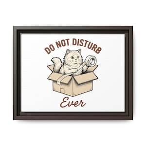 May include: A framed wall art print featuring a fluffy cat sitting inside a cardboard box, holding a roll of adhesive tape. The text DO NOT DISTURB is arched above the cat, and the word Ever is printed below the box in brown lettering.
