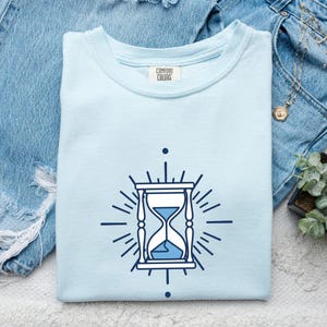 Może przedstawiać: Jasnoniebieski t-shirt z ciemnoniebieską grafiką klepsydry umieszczoną na środku klatki piersiowej. Wzór klepsydry zawiera promieniujące linie oraz kropkę nad i pod centralną ikoną. Wewnętrzna metka na karku zawiera napis Comfort Colors.