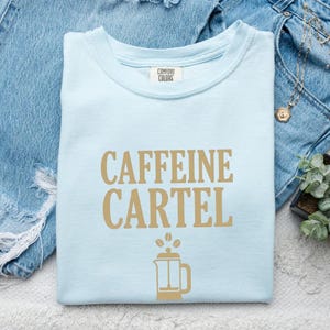 May include: A light blue crew neck t-shirt with the text CAFFEINE CARTEL printed in tan letters. Below the text is a tan graphic of a French press coffee maker with three coffee beans floating above it. The shirt has a Comfort Colors brand label.