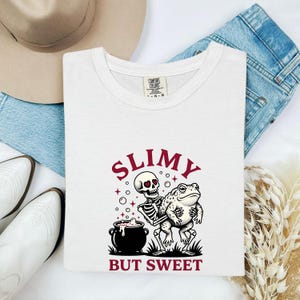 Puede incluir: Una camiseta blanca con un estampado gráfico de un esqueleto con ojos en forma de corazón sosteniendo un sapo junto a un caldero negro burbujeante. El texto sobre y debajo de la ilustración dice SLIMY BUT SWEET en una fuente serif roja oscura.