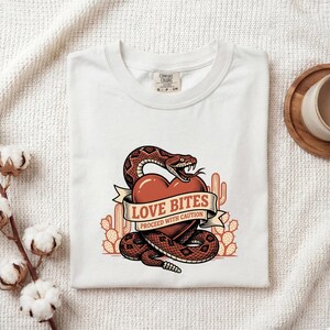 May include: A white t-shirt featuring a graphic of a brown rattlesnake coiled around a red heart. A banner across the heart reads LOVE BITES PROCEED WITH CAUTION. The design includes orange cactus silhouettes in the background.