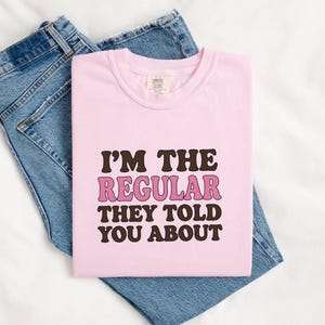 Può includere: Una t-shirt a maniche corte rosa chiaro con il testo marrone scuro I'M THE e YOU ABOUT, e la parola REGULAR in rosa con un contorno marrone scuro. La maglietta è piegata e posizionata sopra un paio di jeans in denim blu.
