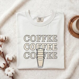 Puede incluir: Una camiseta blanca de manga corta con la palabra COFFEE impresa tres veces en una fuente de contorno negro en negrita. El centro del diseño incluye un gráfico de tres vasos apilados llenos de un líquido beige claro, reemplazando las letras centrales.