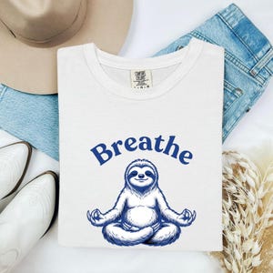 May include: A white short-sleeve t-shirt featuring a blue graphic of a sloth sitting in a lotus meditation pose. Above the sloth, the word "Breathe" is printed in a bold blue font. The shirt has a Comfort Colors brand label at the neck.