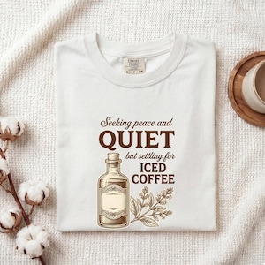 May include: A white short-sleeved t-shirt featuring a brown graphic of a glass bottle and a leafy sprig. The text reads: Seeking peace and QUIET but settling for ICED COFFEE. The shirt has a crew neckline and a Comfort Colors brand label at the neck.