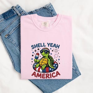 Pode incluir: Uma t-shirt rosa claro com o gráfico de uma tartaruga verde usando óculos de sol em forma de estrela e uma faixa listrada, flutuando numa boia vermelha, branca e azul com um gelado, e o texto SHELL YEAH AMERICA.