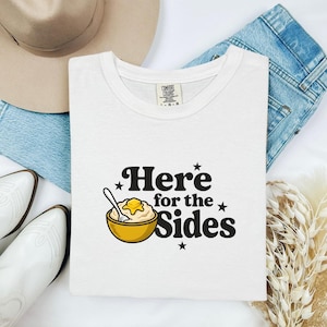 Può includere: Una t-shirt bianca con la grafica di una ciotola gialla piena di purè di patate, guarnita con un fiocchetto di burro e un cucchiaio. Il testo "Here for the Sides" è scritto in nero, circondato da piccole stelle nere come elemento decorativo.