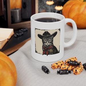 May include: A white ceramic mug featuring a graphic illustration of a cat wearing a cowboy hat, a red neckerchief, and a sheriff star badge. The cat is depicted in black and white ink style against a square beige background with a distressed border.