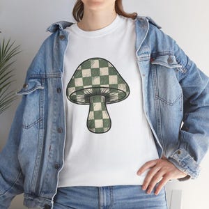 May include: A white t-shirt featuring a large graphic of a mushroom with a green and cream checkered pattern on the cap and stem. The mushroom design includes fine black line work and shading. The shirt is worn under a light blue denim jacket.