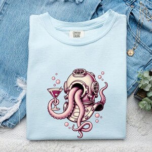 May include: A light blue t-shirt featuring a graphic of a vintage diving helmet with pink octopus tentacles emerging from the faceplate. One tentacle holds a martini glass with a red olive. Small pink bubbles surround the helmet design.