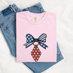 Op de afbeelding: Een lichtroze t-shirt met een afbeelding van een patriottische strik met blauwe, witte en rode sterren- en strepenpatronen, bevestigd aan een gouden ring met een rode ruitvormige hanger met witte sterren, gecombineerd met een blauwe spijkerbroek.