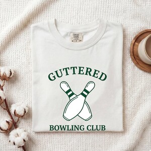 May include: A white crew neck t-shirt featuring a dark green graphic of two crossed bowling pins. The text "GUTTERED" is printed in a dark green arched font above the pins, and "BOWLING CLUB" is printed in dark green below the pins.