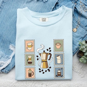 Puede incluir: Una camiseta azul claro con un gráfico central de una cafetera italiana rodeada de granos de café y seis ilustraciones rectangulares estilo sello. Estos sellos muestran diversos equipos de preparación de café en tonos apagados de beige, verde y azul.