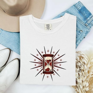 May include: A white t-shirt featuring a graphic of a brown hourglass surrounded by radiating dark red lines and small star shapes. The shirt has a Comfort Colors label at the inner neckline. The design is centered on the chest of the garment.