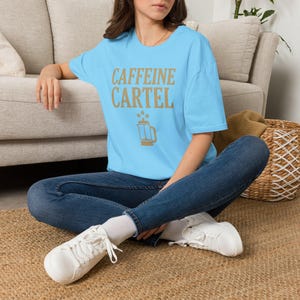 May include: A light blue short-sleeve t-shirt featuring the text CAFFEINE CARTEL in gold lettering, positioned above a gold graphic of a French press coffee maker with three coffee beans floating above it, worn with dark blue denim jeans.