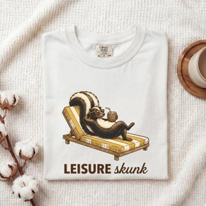 Könnte beinhalten: Ein weißes T-Shirt mit einer Grafik eines Stinktiers mit Sonnenbrille, das auf einer gelb-weiß gestreiften Liege entspannt und ein Ananasgetränk hält. Der Text LEISURE skunk ist in Braun unter der Illustration aufgedruckt.