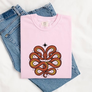Op de afbeelding: Een lichtroze t-shirt met een afbeelding van een opgerolde oranje slang met een gele buik, gerangschikt in een symmetrisch patroon met een zwarte vierpuntige ster erboven, gecombineerd met een blauwe spijkerbroek.