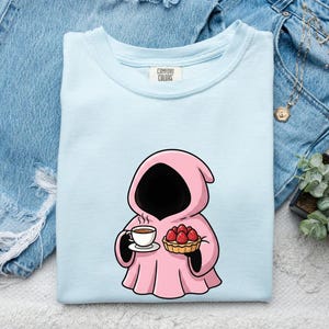 May include: A light blue t-shirt featuring a graphic of a pink hooded figure holding a white teacup and a strawberry tart. The inner neck label displays the text Comfort Colors. The shirt is folded and presented on a textured white surface.