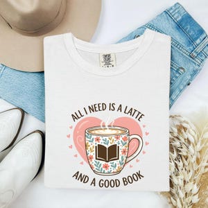 Può includere: Una t-shirt bianca con la grafica di una tazza da caffè floreale con l'icona di un libro aperto, centrata sopra un cuore rosa. Il testo recita All I Need Is A Latte And A Good Book. La maglietta ha un'etichetta Comfort Colors sul collo.