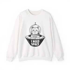 May include: A white long-sleeve sweatshirt featuring a black and white graphic of a cat inside a flying saucer. The cat wears a glass helmet, and the text "I NEED SPACE" is printed in bold white capital letters on the side of the spacecraft.