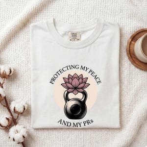 May include: A white short-sleeved t-shirt featuring a graphic of a black kettlebell topped with a pink lotus flower. The text "PROTECTING MY PEACE AND MY PRs" is printed in a circular arc around the central illustration in black lettering.