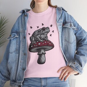 May include: A light pink t-shirt featuring a black and white graphic illustration of a toad sitting on a large red mushroom with white spots, surrounded by small flying insects. The t-shirt is worn under a light blue denim jacket with metal buttons.