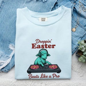 May include: A light blue t-shirt featuring a graphic of a teal lamb wearing headphones and operating a DJ turntable. The text above the lamb reads "Droppin' Easter" and the text below reads "Beats Like a Pro" in a dark red font.