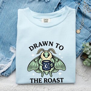 Può includere: Una t-shirt azzurra con la grafica di una falena che tiene in mano una tazza blu scuro con un disegno di luna e stelle. Il testo sopra la falena recita DRAWN TO e il testo sotto recita THE ROAST. La maglietta ha un'etichetta Comfort Colors.